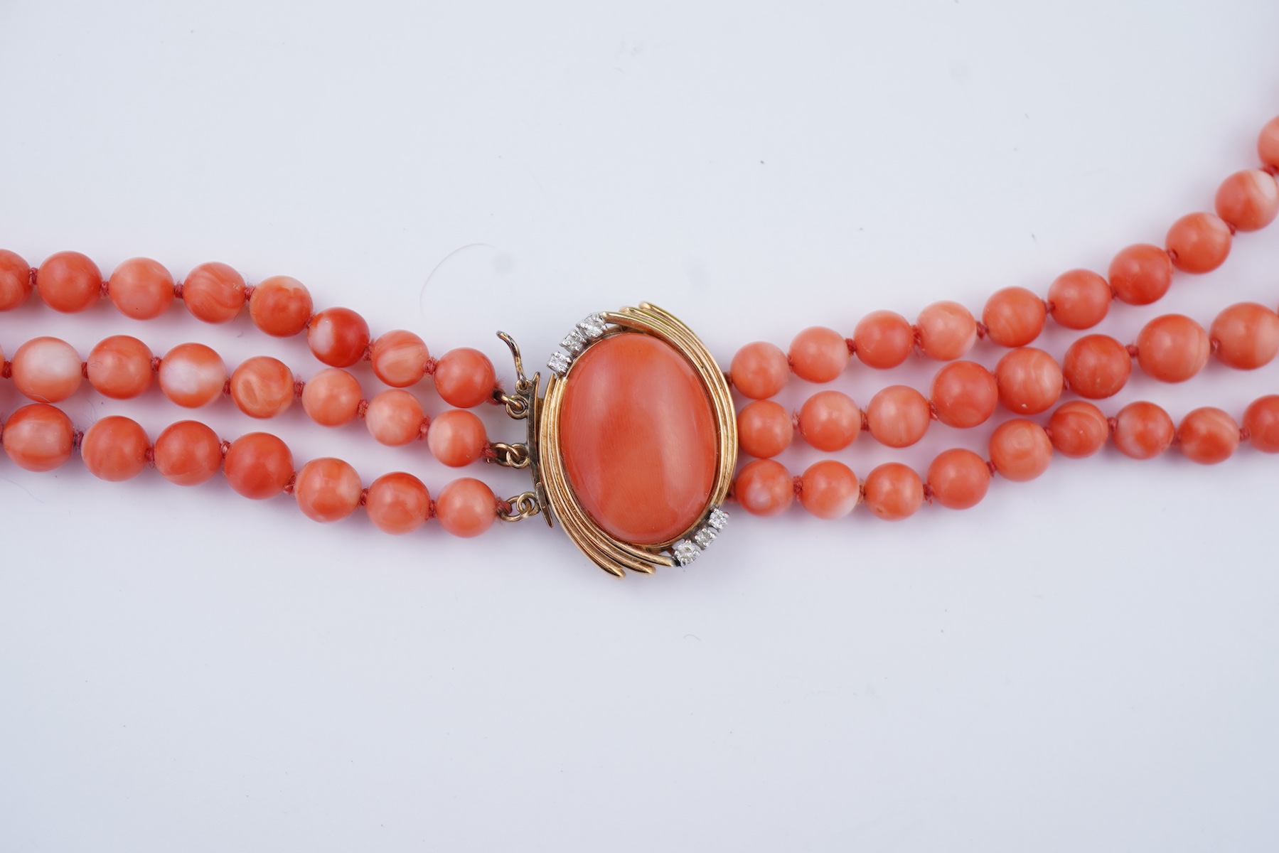 A coral necklace, composed of three strands of coral beads measuring approximately 5.1-6.6mm diameter, to a gold clasp set with a coral cabochon and brilliant-cut diamonds, mounted in gold, length 45.5cm, stamped 14K, ca